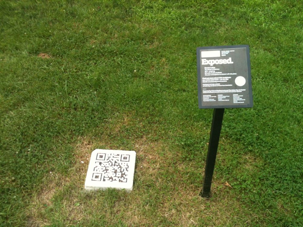 A rectangular cube plaster mold that resembles a QR code on a grassy field with a sign that says 'Exposed' and includes additional information about the project