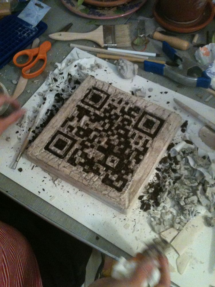 A rectangular cube plaster mold that contains emptied cavities that resemble the outlines of a QR code filled with dirt on a work table with scissors and brushes and other tools scattered on the table