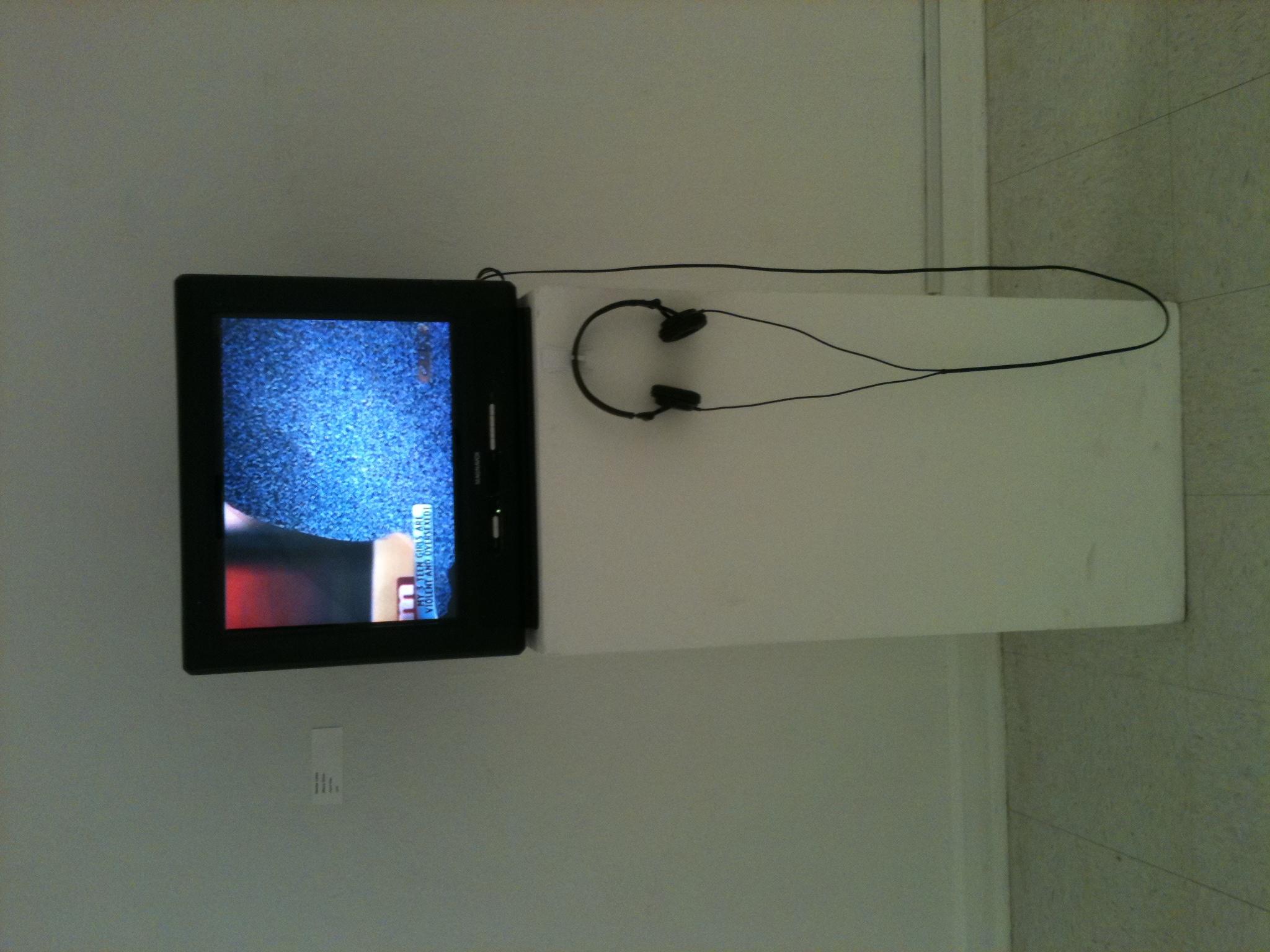 A TV monitor is on a pedestal. The image on the screen is a silhouette of a person sitting on a couch and TV static fills the figure. Headphones hang on the pedestal.